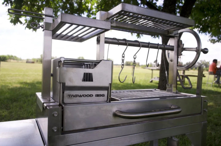 Tagwood Freestanding "Mini" Santa Maria Style Grill: Compact stainless steel grill with four distinct cooking zones, featuring height-adjustable main grate, interchangeable griddle, secondary grate, and S-shape hooks for hanging meat.