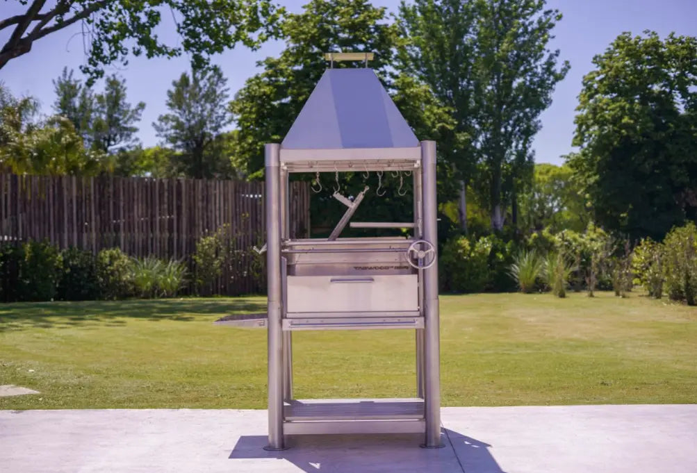 Tagwood "The Silver Bullet" Santa Maria Style Grill - Stainless Steel: A freestanding, stainless steel grill with 1,700 sq. in. cooking area, six surfaces, height-adjustable grates, and firebrick insulation for authentic 360° open-fire experience.