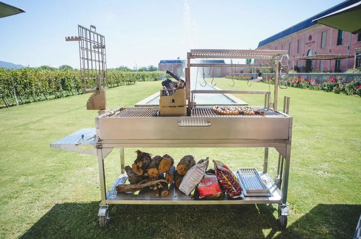 Tagwood XL Freestanding Argentine Santa Maria Style Grill: Stainless steel grill station with five cooking zones, adjustable grates, and firebrick-lined walls for even heat distribution, featuring 2,600 sq. in. of total grilling area, foldable side table, and heavy-duty casters for mobility.