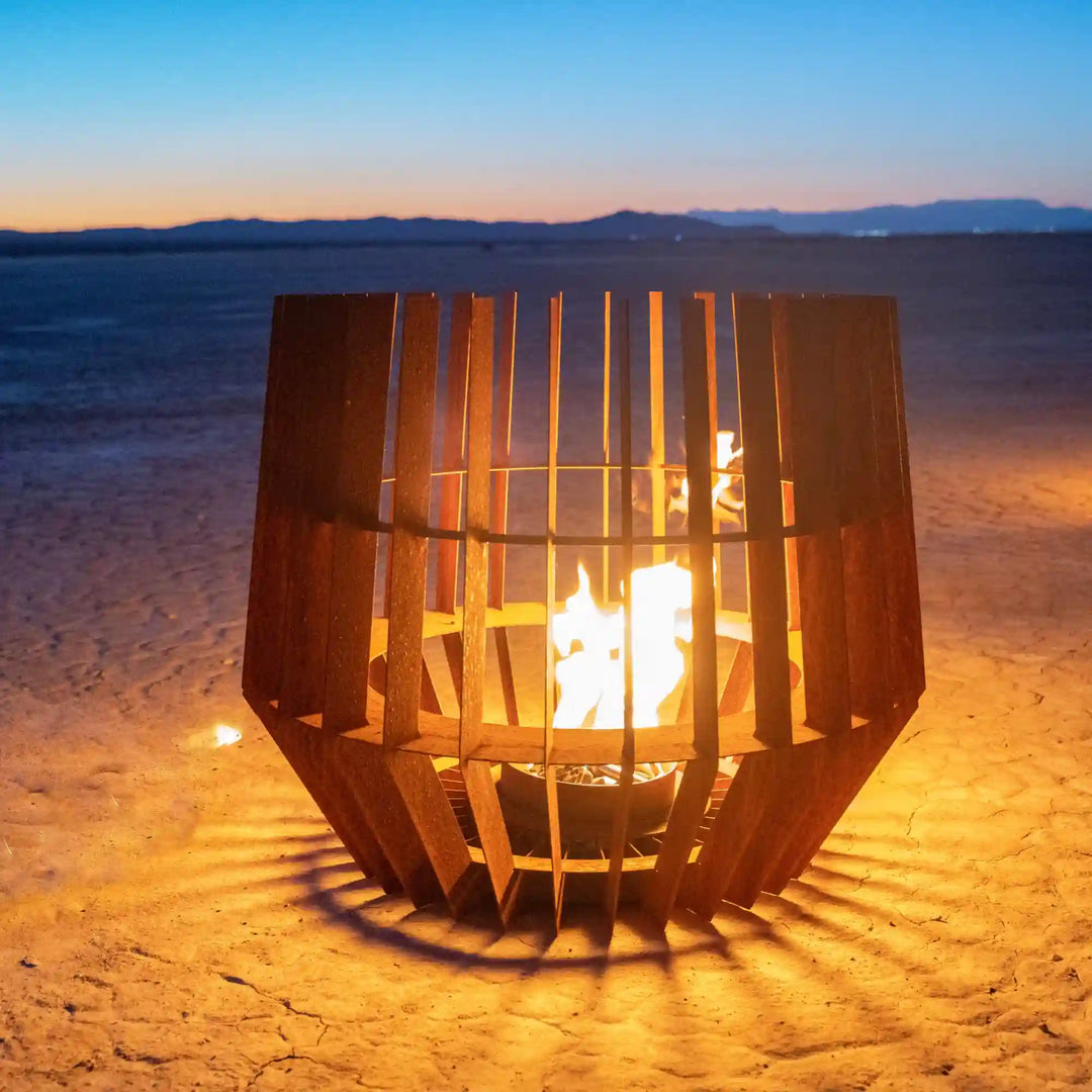 Sol Fire Sculpture - The Outdoor Plus: A modern, tall, open flame sculpture made from vertical Corten steel panels forming a cage, showcasing a bright glow through slats in a warm and bold outdoor centerpiece.