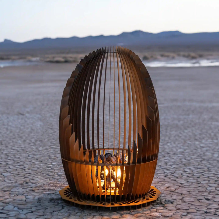 Nebula Fire Sculpture - The Outdoor Plus: A modern, oval-shaped fire feature made from vertical Corten steel slats wrapped around a radiant flame, emitting warmth and visual appeal.