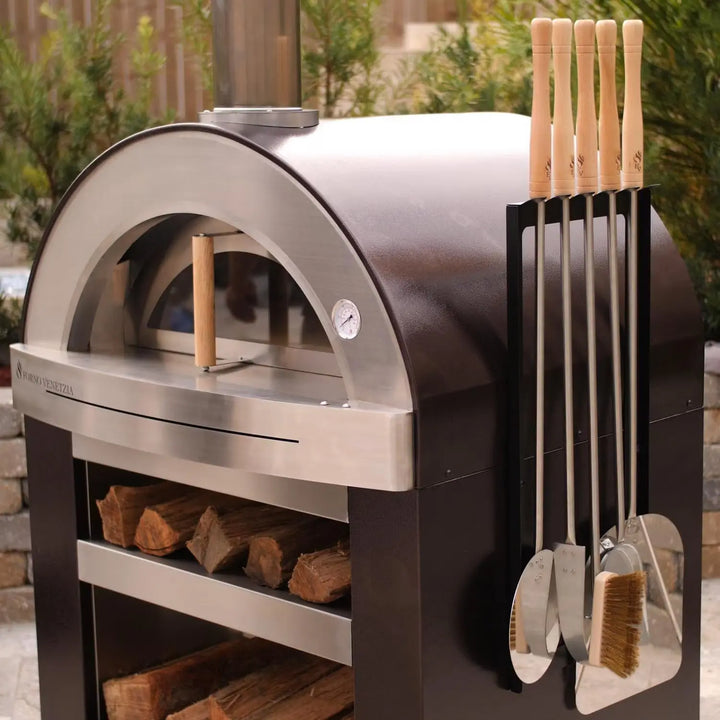 The Forno Venetzia "Torino 500" - Copper outdoor pizza oven features a spacious 32" × 24" cooking surface (768 sq. in.) and a firebrick hearth for authentic flavor, complemented by a stainless steel cart with pull-out side shelf and adjustable tool holder, finished in UV-resistant copper dome with signature chimney cap and oak handle.